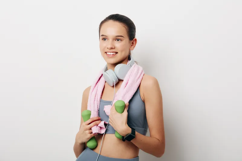 Female fitness enthusiast holding green dumbbells, pink towel draped around her neck, wireless headphones, gray sports top, mid chest pose in a clean studio backdrop, ready for exercise and training.