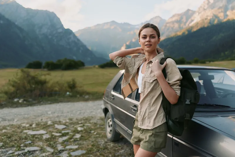 A young woman backpacker stands beside a dark car in a sweeping mountain landscape, adjusting her backpack straps as she prepares for a hiking adventure, while distant peaks loom.