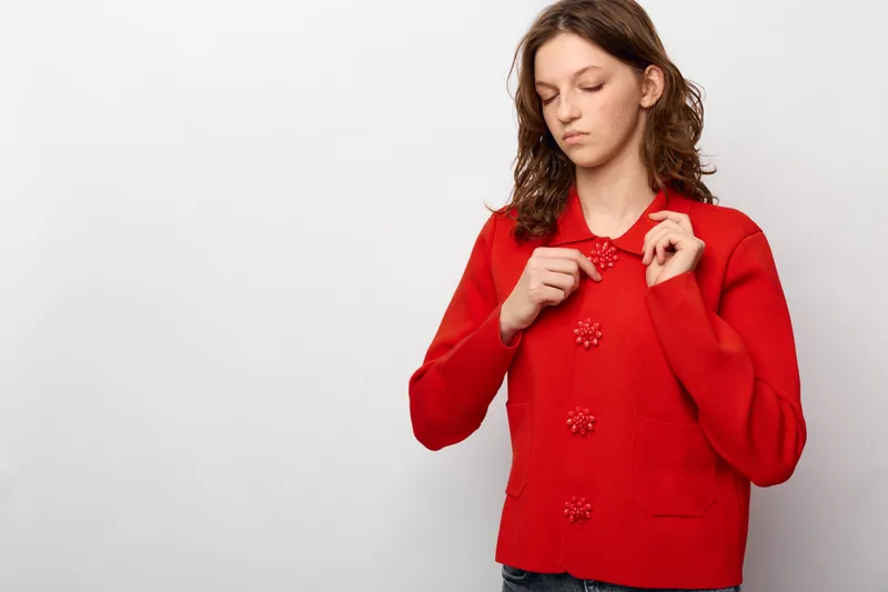 Young woman in red knitted sweater buttoning up with focused expression on white background. Casual outfit, autumn fashion, and cozy clothing concept with natural light and simple studio setting.