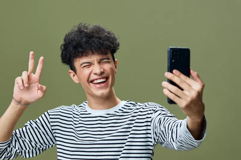 smiling man taking selfie with smartphone wearing striped shirt showing peace sign on olive green background, joyful expression, casual style