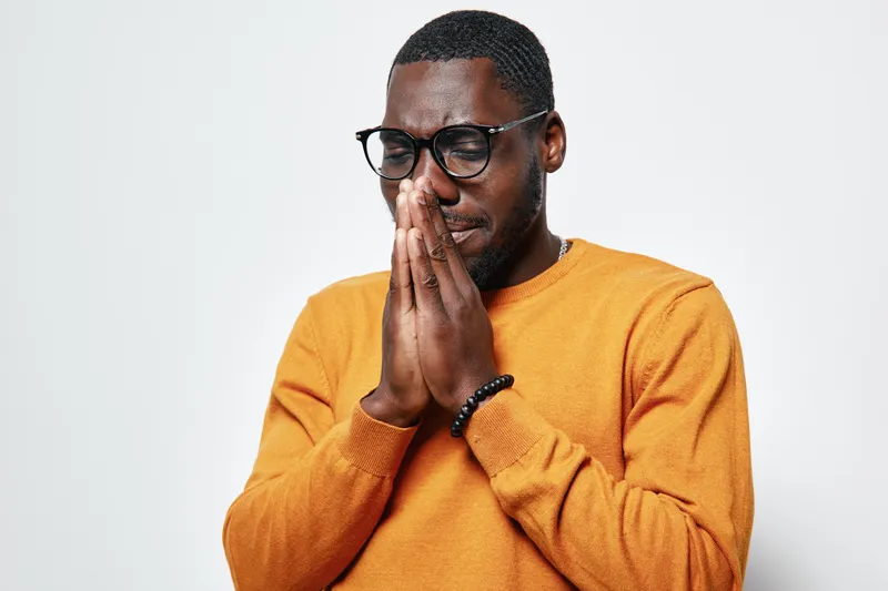 African American man with dark skin and glasses wearing orange sweater praying with closed eyes and hands together isolated on white background for faith and hope concept.