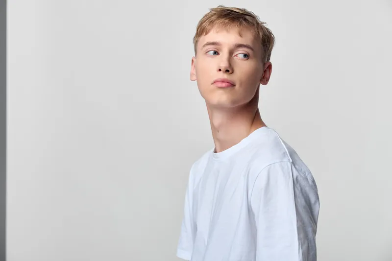 Young man in a white shirt turns slightly away against a neutral studio backdrop, presenting casual style, a thoughtful expression and modern, calm appeal