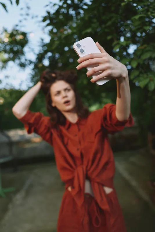 Woman lifestyle scene outdoors in a red dress, taking a selfie with a smartphone, capturing a cheerful, carefree moment against a leafy backdrop and a soft daylight glow