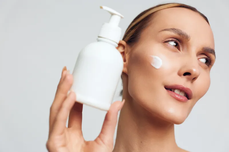 Bright woman applying moisturizer, close up of face with a pump bottle, soft studio lighting, dewy skin, calm expression, skincare routine, hydration, healthy glow, minimal background