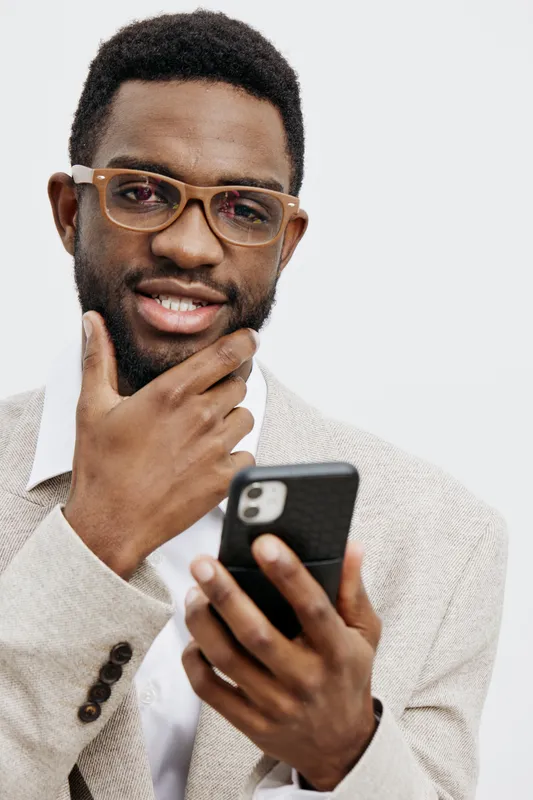 Stylish young African American man wearing glasses and a beige blazer holding a smartphone, thinking and smiling, isolated on a plain white background. People lifestyle concept