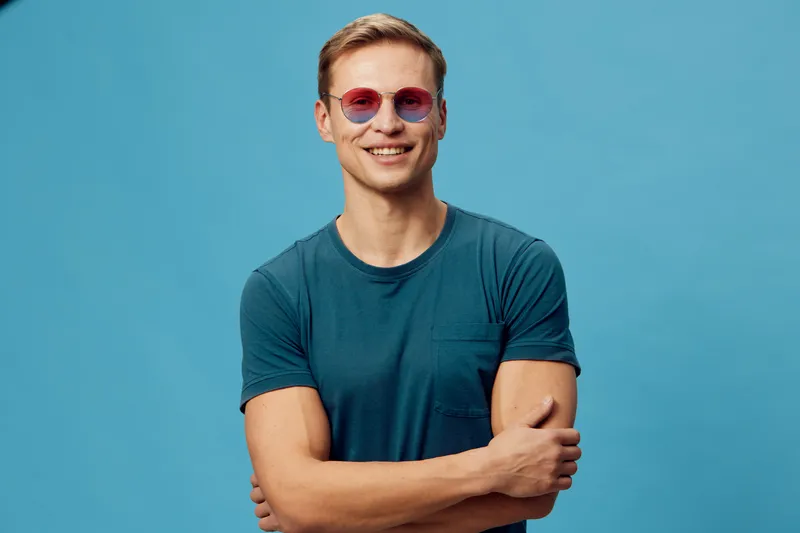 Smiling young man with short hair wearing pink sunglasses and casual teal t-shirt stands confidently against a plain blue background, looking directly at the camera. Portrait of cheerful male model, f