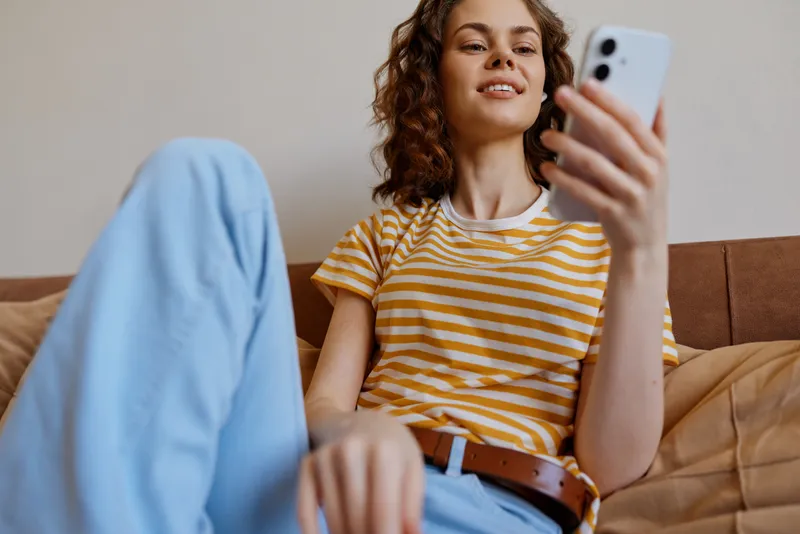 A woman on a sofa at home captures a casual selfie in a striped tee and blue jeans, showcasing a relaxed lifestyle moment and everyday comfort and self expression.