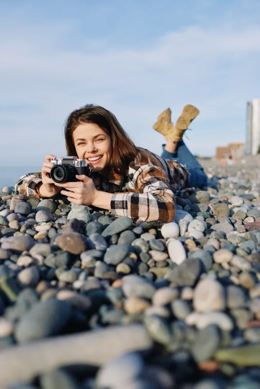 Photographer lying among beach pebbles, smiling while filming with a camera, outdoor lifestyle scene on a sunny shore, candid creative moment captured among rocks and open sky