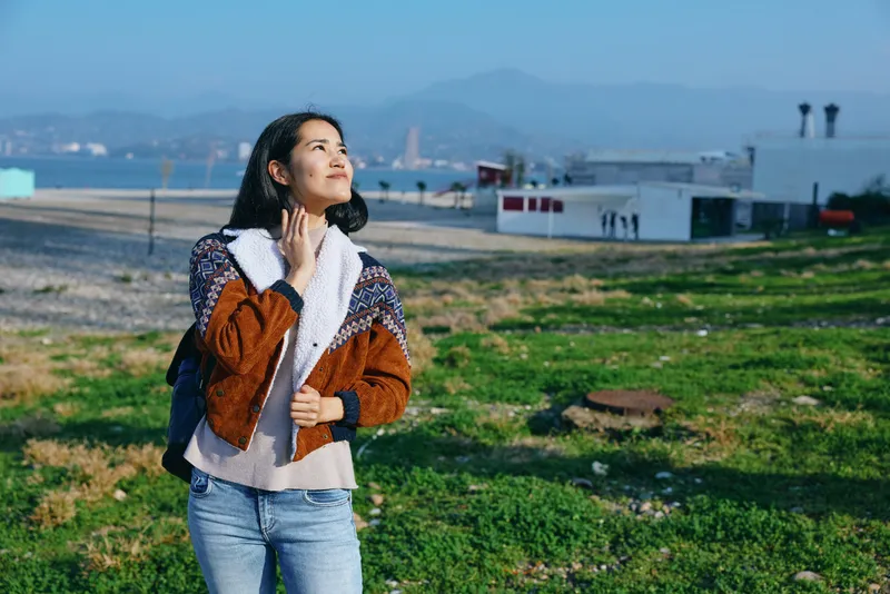 Woman in a stylish coat and scarf stands outdoors on a grassy field looking up with confidence urban rooftops in distance warm natural light casual fashion vibe