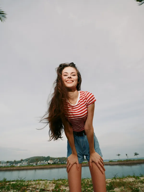 A cheerful young woman stands outdoors in a bright striped shirt, smiling warmly in the sun with a carefree pose that conveys energy, youth, and natural joy.