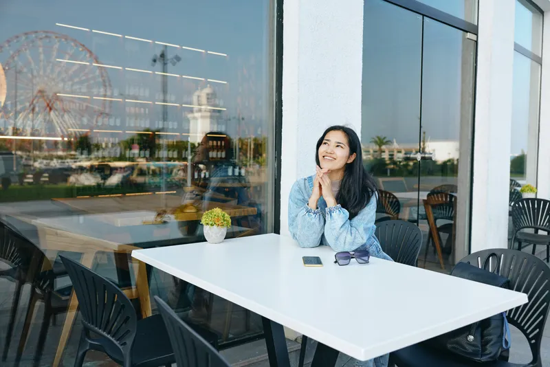 Woman sits at a white table at an outdoor cafe, hands folded under chin, bright day, reflective sunglasses, potted plant, glass windows, urban calm, casual dining mood.
