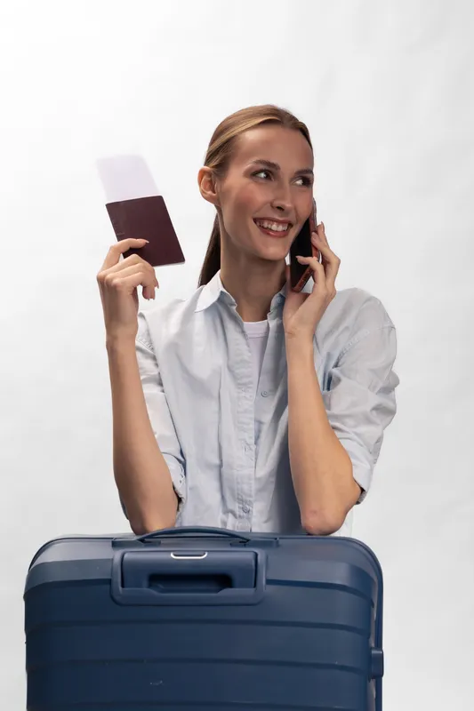 Young woman holding passport and boarding pass talks on phone, standing behind blue suitcase. Concept for travel planning, vacation, and communication.