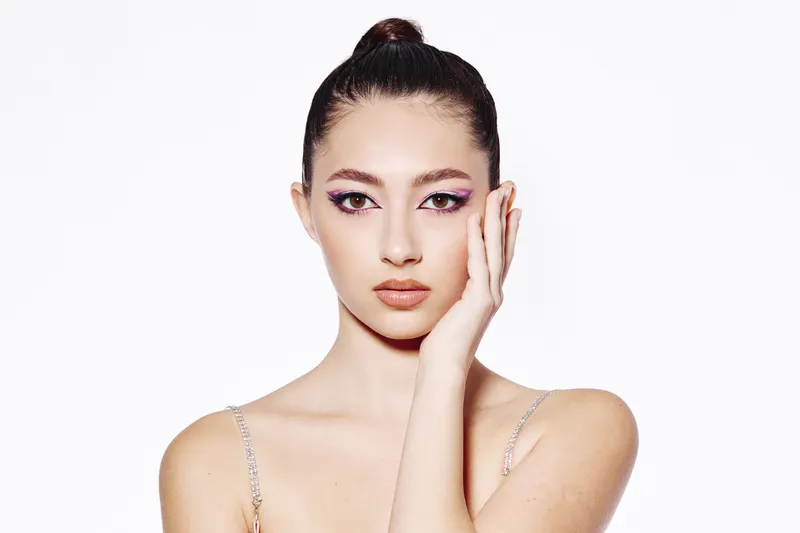 Young woman with creative purple luminous eye makeup touches face against a pure white background. Concept for beauty, fashion, creative style and modern grooming.