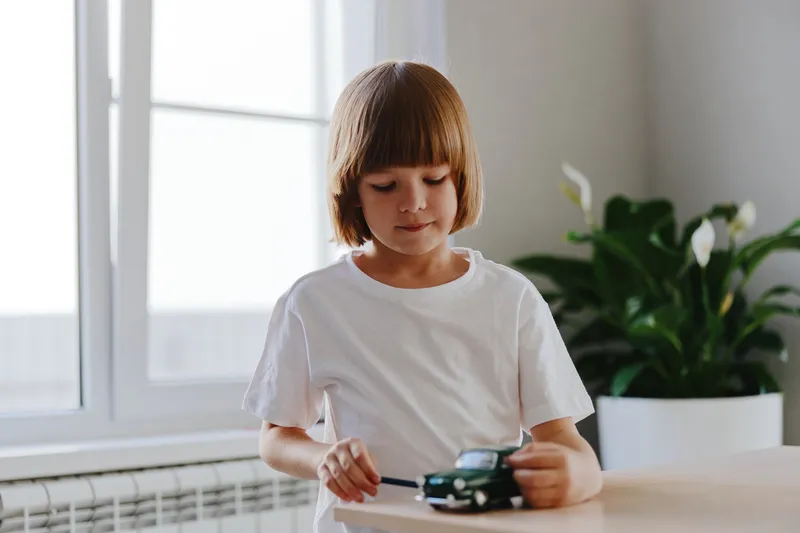 A young girl enjoys playing with a toy car on a wooden table, surrounded by plants in a bright and cozy room. Her focused expression captures the joy of creativity.