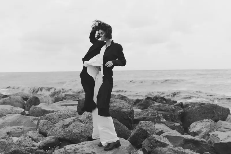 Dancer swirls on a rocky shoreline by the sea, flowing garments captured in monochrome as wind lifts fabric and conveys motion and freedom