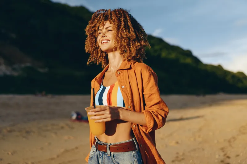 Woman on beach smiling outdoors with curly hair in warm sunlight, brown jacket and denim, striped top, relaxed stance on sandy shore, distant hills under evening sky, gentle breeze and sea