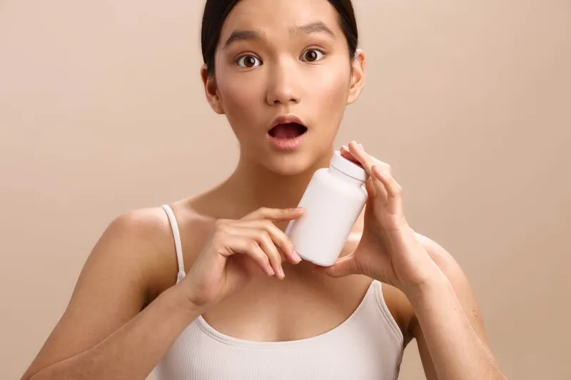 asian woman holds a white bottle up to her mouth, eyes wide in a soft, neutral setting. she appears curious and amused, creating a playful and engaging mood.