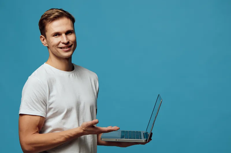 Friendly smiling young man with brown hair wearing casual white t-shirt holding a laptop in hand, standing against a plain blue background. Studio portrait with a positive and approachable demeanor. P