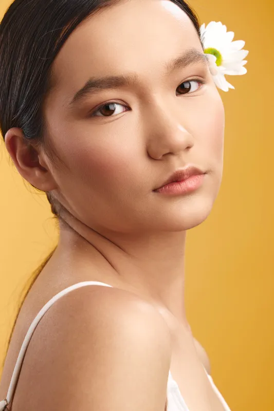 asian woman with a delicate glow stands against a warm yellow backdrop, a white strap top reveals elegant shoulders, soft lighting enhances natural beauty and serene mood.