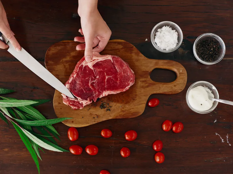 Женщина с ножом в руке нарезает свежее мясо для стейка для запекания на кухне