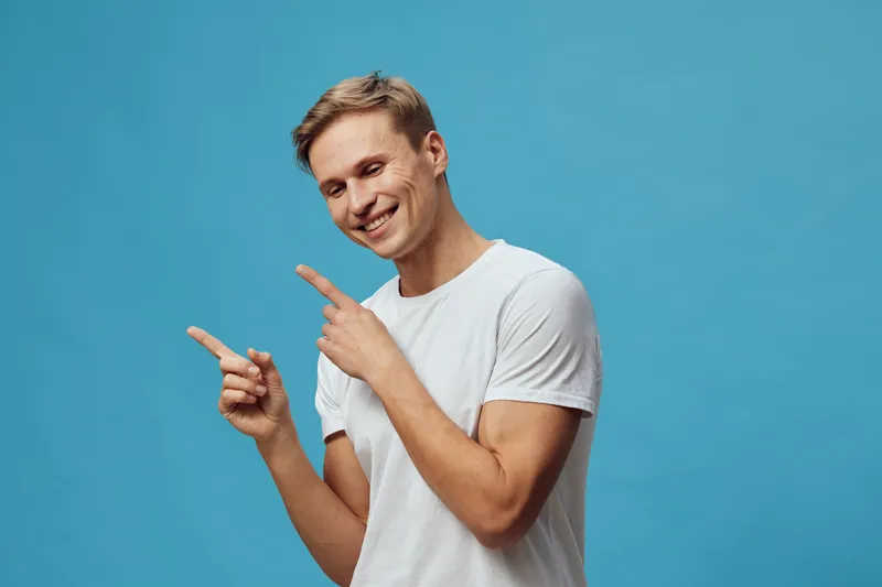 Happy smiling man pointing with fingers, casual white t-shirt, light brown hair, cheerful expression, isolated on blue background, studio portrait, positive emotion, lifestyle concept, young adult mal