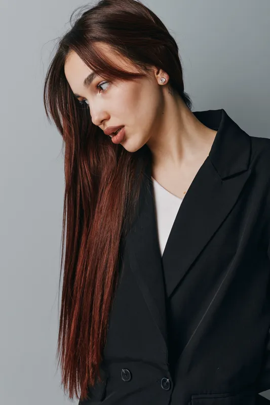 Woman with long straight hair and light skin wearing black blazer and white shirt posing against gray background with soft natural makeup and earrings in studio portrait.