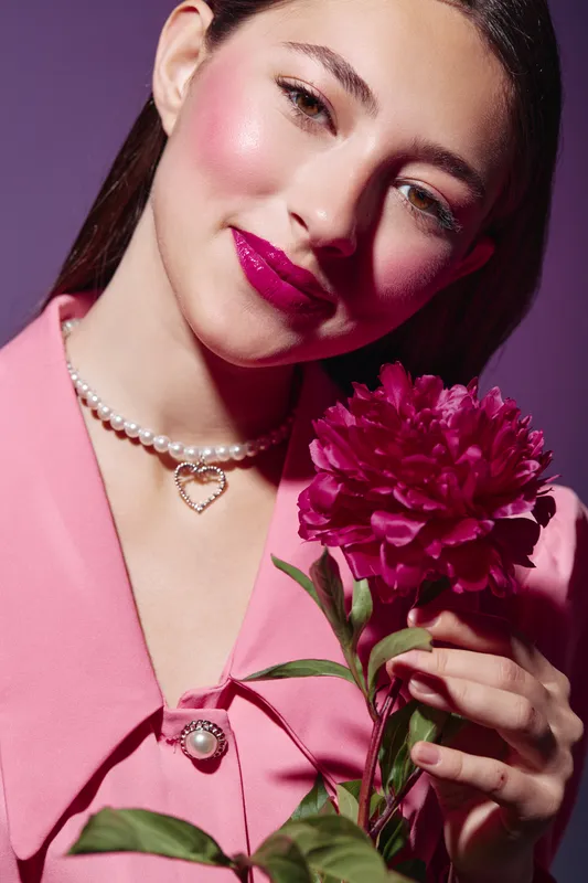 Young woman with vibrant pink makeup smiles gently holding bright pink flower. Fashion style with glossy lips, glowing skin, and pearl necklace. Studio background concept for beauty.
