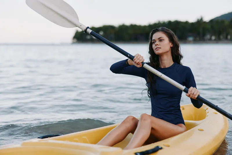 Young woman paddling a yellow kayak on calm water, wearing a dark blue wetsuit, focused expression, wind in hair, with scenic shoreline visible in the distance.