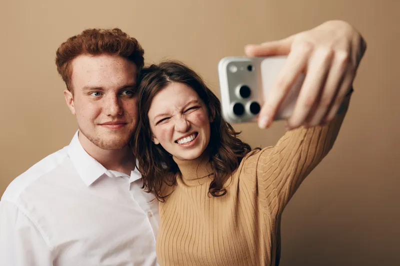 Young couple takes a selfie together, smiling and posing in casual clothes against a plain beige background. Concept of friendship, happiness, and modern lifestyle with smartphone technology.
