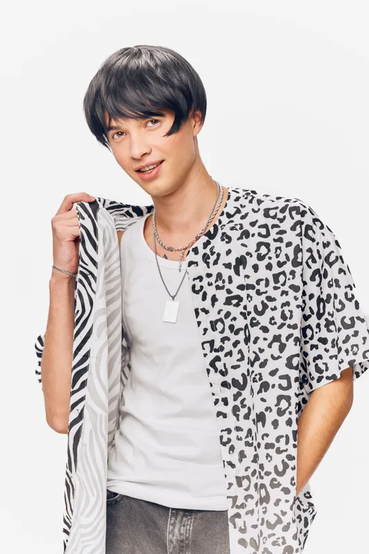 young man wearing patterned black and white shirt holds matching fabric while smiling confidently against white background, casual style with modern accessories