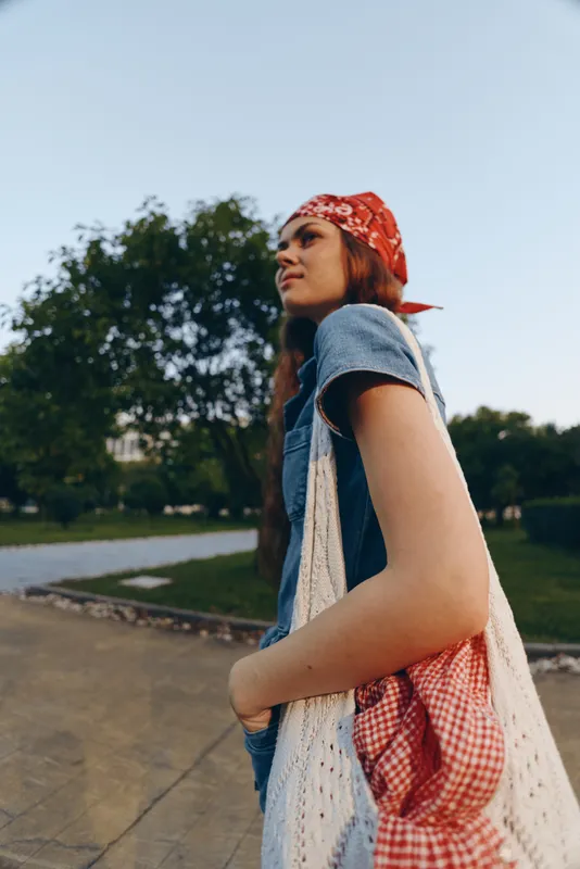 young woman with red bandana walking outdoors in casual denim outfit carrying a tote bag in a green park with clear sky during daytime