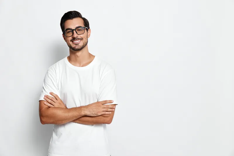 Confident smiling young man wearing glasses and a casual white t-shirt, posing with crossed arms in front of a light gray wall. Modern lifestyle concept.