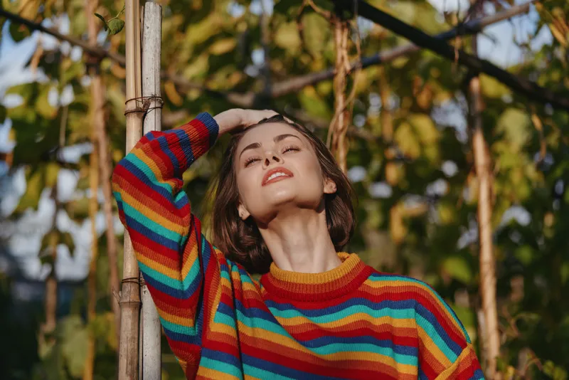 Woman in striped sweater leaning on bamboo outdoors, young adult smiling with eyes closed in tropical garden, casual outfit, relaxed pose and warm sunlight for lifestyle or fashion travel.