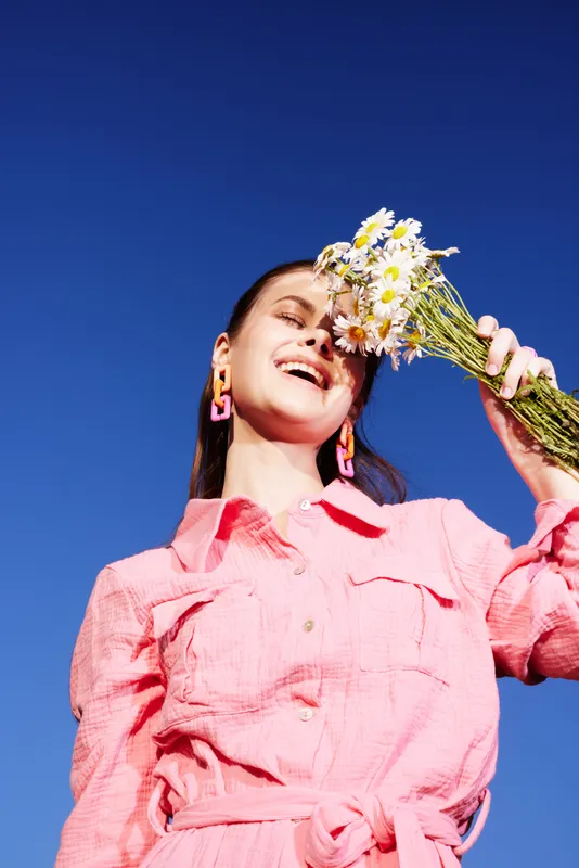 Joyful woman in pink outfit holding daisies under a clear blue sky, radiating happiness and freshness with vibrant colors and summer vibes