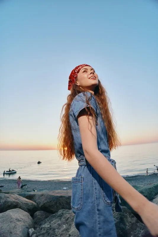 lifestyle woman in modern boho-western denim street style with red bandana, long curly hair, and sunset beach background in warm film color tones