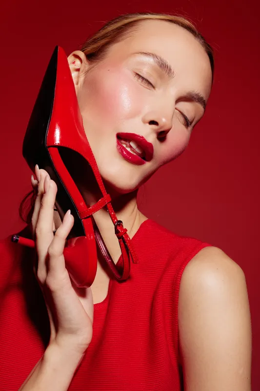 Woman with closed eyes holds red high heel shoe near face, wearing red sleeveless top. Glossy makeup and shiny skin on red background. Concept of fashion, beauty, and style.