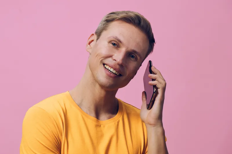 Bright young man with short blond hair smiling and talking on smartphone against pink background, casual yellow t-shirt, lifestyle, communication concept.