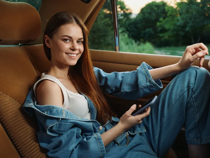 Young woman in casual denim outfit sitting in a car, smiling and using a smartphone, surrounded by nature in the background, evoking a sense of freedom and joy.
