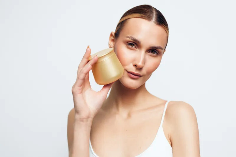 A young woman with clear skin holds a jar of cream near her face against a white background. Concept for skincare, beauty, natural cosmetics, and personal care routines.