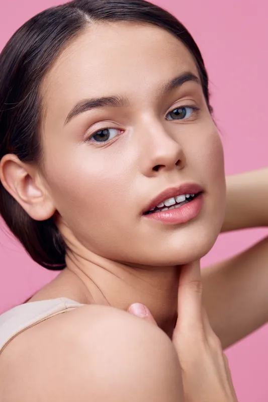 Youthful female model with natural makeup and soft expression against a pink background, showcasing beauty and modern skincare trends
