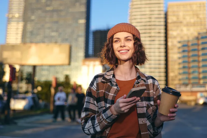woman holding coffee and phone in city, smile and urban vibe during golden hour; candid lifestyle portrait capturing authenticity, mindful living and emotional storytelling with warm glow