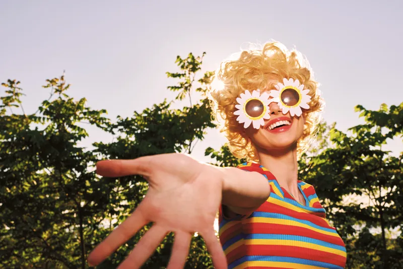 Vibrant retro woman outdoors, wearing a colorful striped top, playful smile, sunlit scene, carefree lifestyle, nature background, daisies on glasses, warm tones