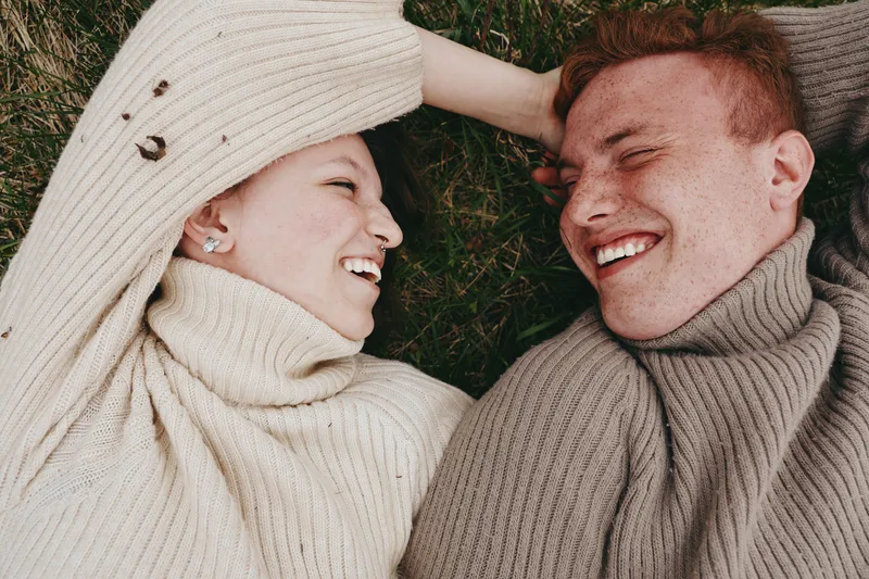 A couple lies on grass wearing cozy sweaters, sharing a warm laugh, relaxed lifestyle moment captured outdoors, intimate connection, candid portrait of happiness and affection.