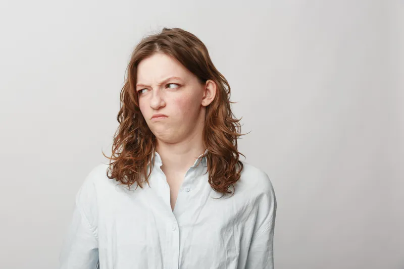 Young woman with curly hair wearing white shirt looks disgusted and skeptical, isolated on light background. Concept of negative emotions, doubt, and displeasure.