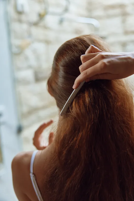 Woman combing long brown hair inside a softly lit room, showing delicate fingers and smooth texture in natural ambiance with warm tones and blurred background.