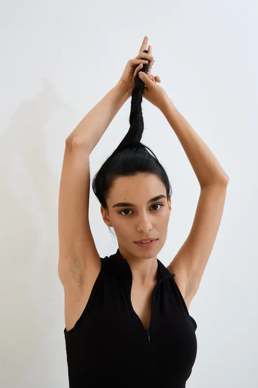 young woman pulling her long black hair upward wearing a sleeveless black top with a calm expression against a plain white background