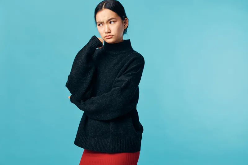 Young woman in black sweater and red skirt standing with thoughtful expression against blue background. Casual clothing, pensive mood and indoor studio portrait of female model.