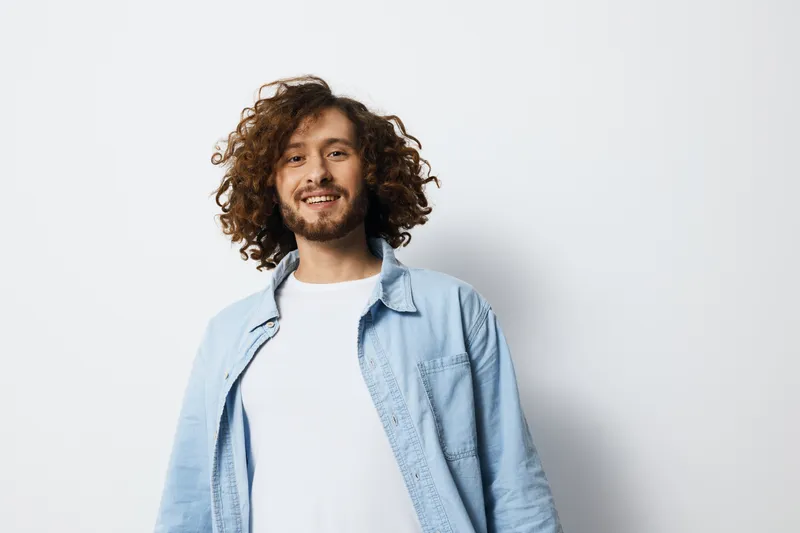 Cheerful young man with curly hair smiling against a plain white background, wearing a light blue shirt Perfect for concepts related to happiness, youth, and positivity