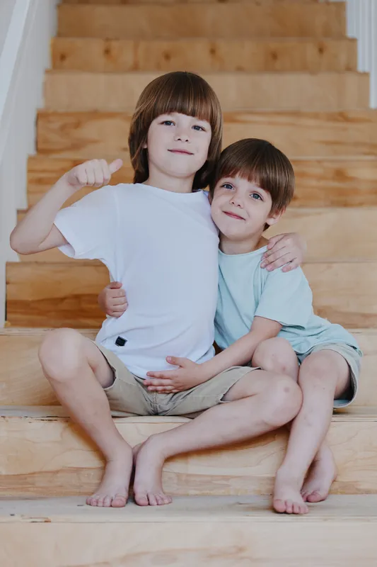 Two happy boys sitting on wooden stairs, joyfully embracing each other with smiles, showcasing friendship and childhood innocence in a warm light.