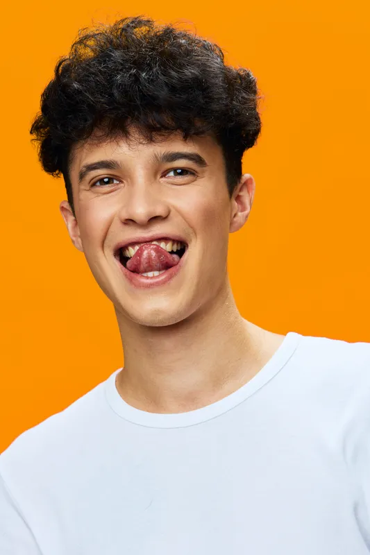 playful young man with curly hair wearing white t-shirt shows tongue against vibrant orange background, expressing joy and fun with energetic smile